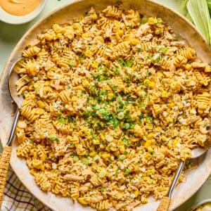 A large dish of Mexican street corn pasta salad mixed with green onions and a creamy orange-colored sauce, garnished with chopped herbs. Slices of corn, sauce, and chopped herbs are placed around the main dish.