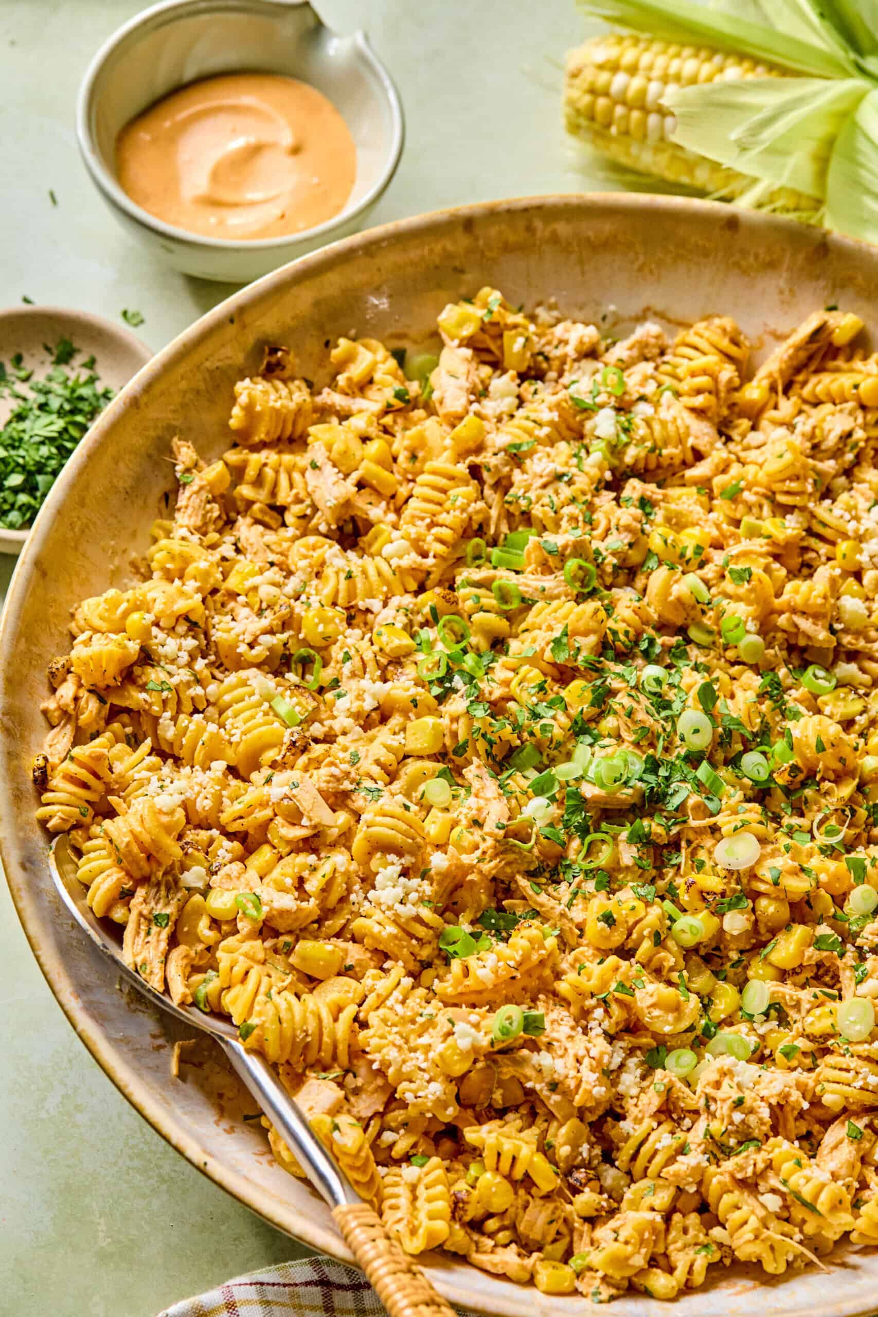 A large bowl of Mexican street corn pasta salad with rotini, corn, green onions, crumbled cheese, and herbs, served with a creamy sauce in a small bowl and fresh corn on the cob in the background.