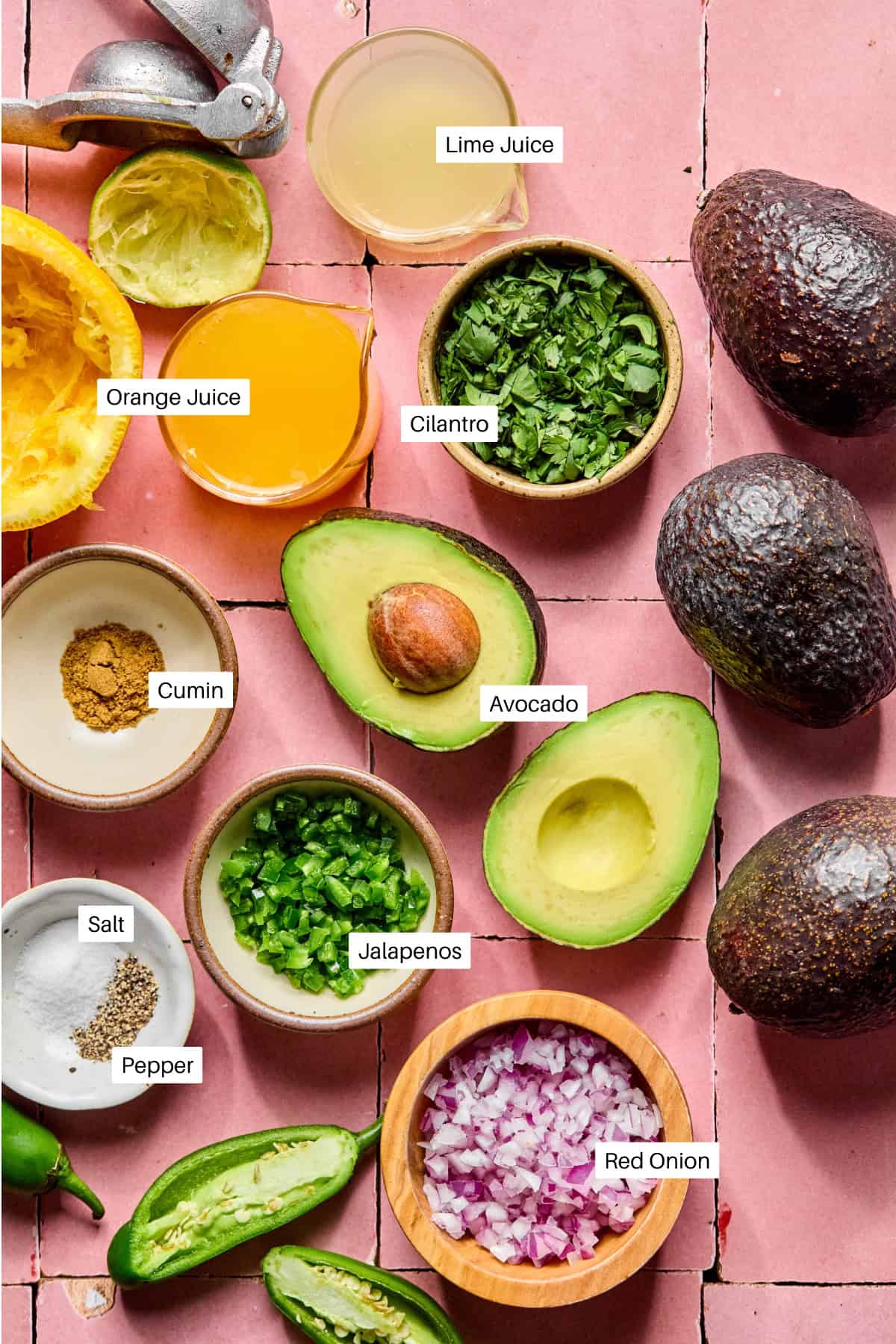 Ingredients for an Easy Guacamole Recipe on a pink tile background, including avocados, cilantro, jalapeños, chopped red onion, orange juice, lime juice, cumin, salt, pepper, and a citrus juicer—each clearly labeled.