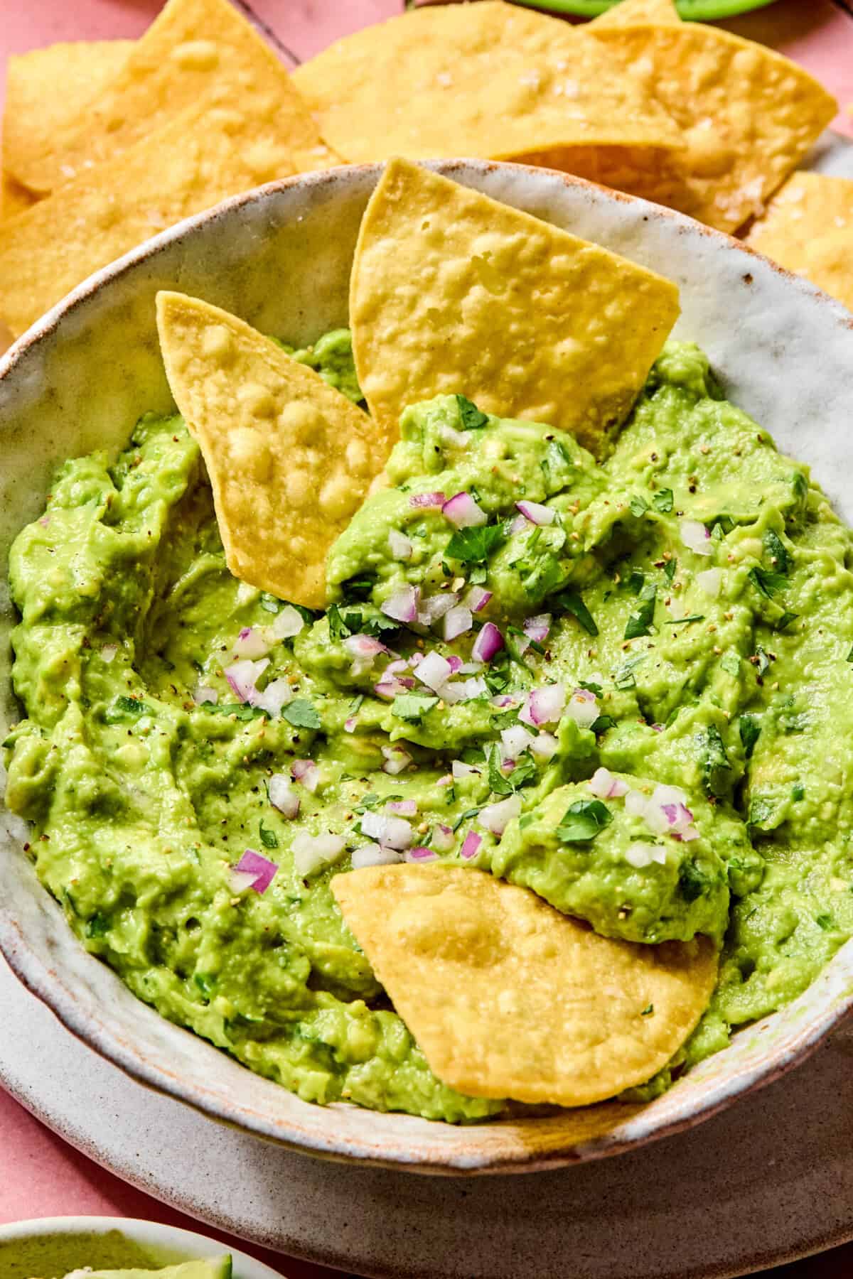 A bowl of Easy Guacamole Recipe topped with chopped red onion and cilantro, served with yellow tortilla chips, some of which are dipped into the creamy guacamole.