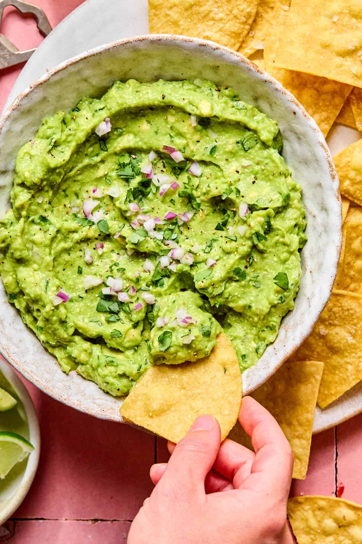 A hand dips a tortilla chip into a bowl of guacamole made from an easy guacamole recipe, topped with chopped red onion and cilantro, surrounded by more chips and lime wedges.