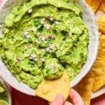 A hand dips a tortilla chip into a bowl of guacamole made from an easy guacamole recipe, topped with chopped red onion and cilantro, surrounded by more chips and lime wedges.