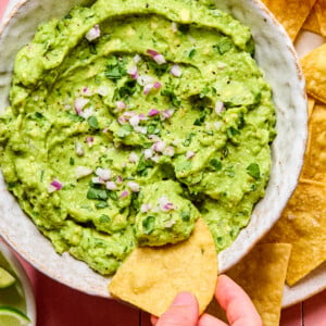 A hand dips a tortilla chip into a bowl of guacamole made from an easy guacamole recipe, topped with chopped red onion and cilantro, surrounded by more chips and lime wedges.