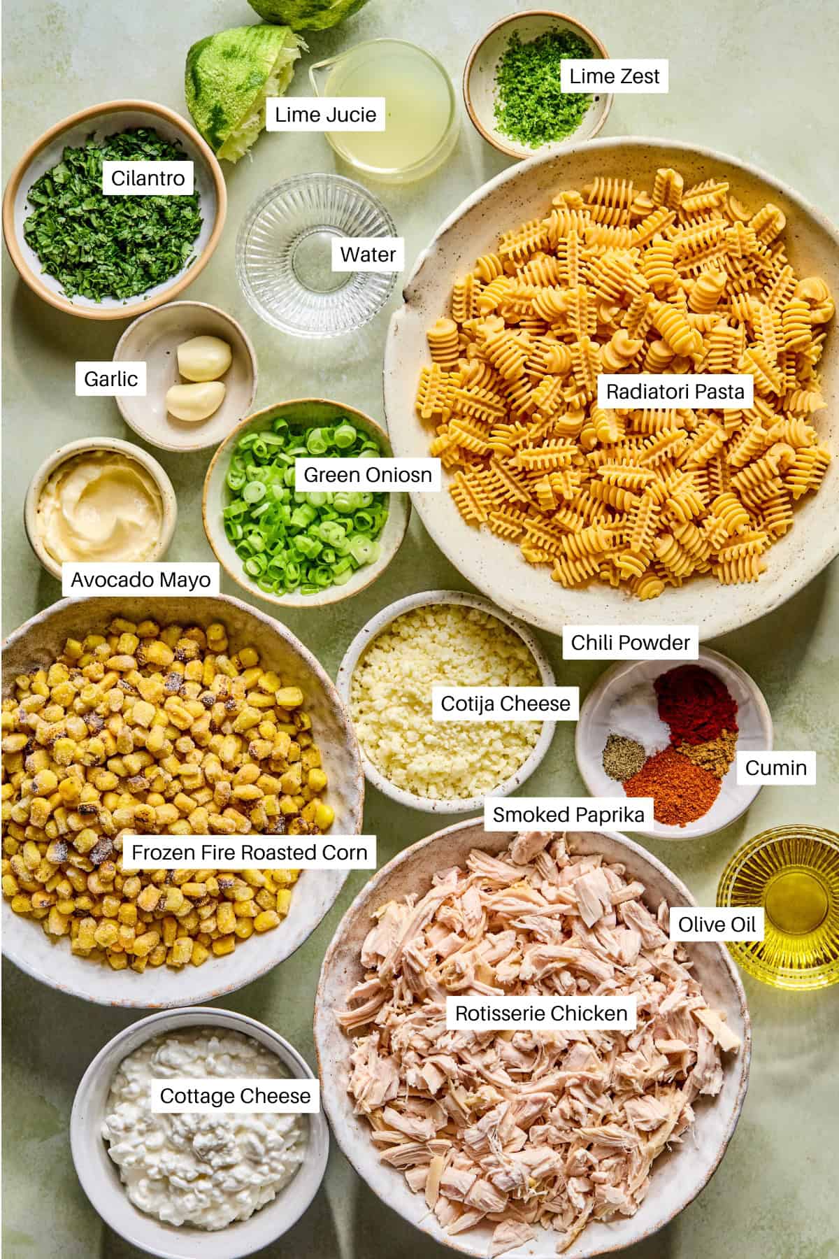Various labeled ingredients for a Mexican street corn pasta salad are arranged on a table, including radiatori pasta, rotisserie chicken, fire roasted corn, avocado mayo, cilantro, green onion, spices, cheeses, lime zest, and lime juice.