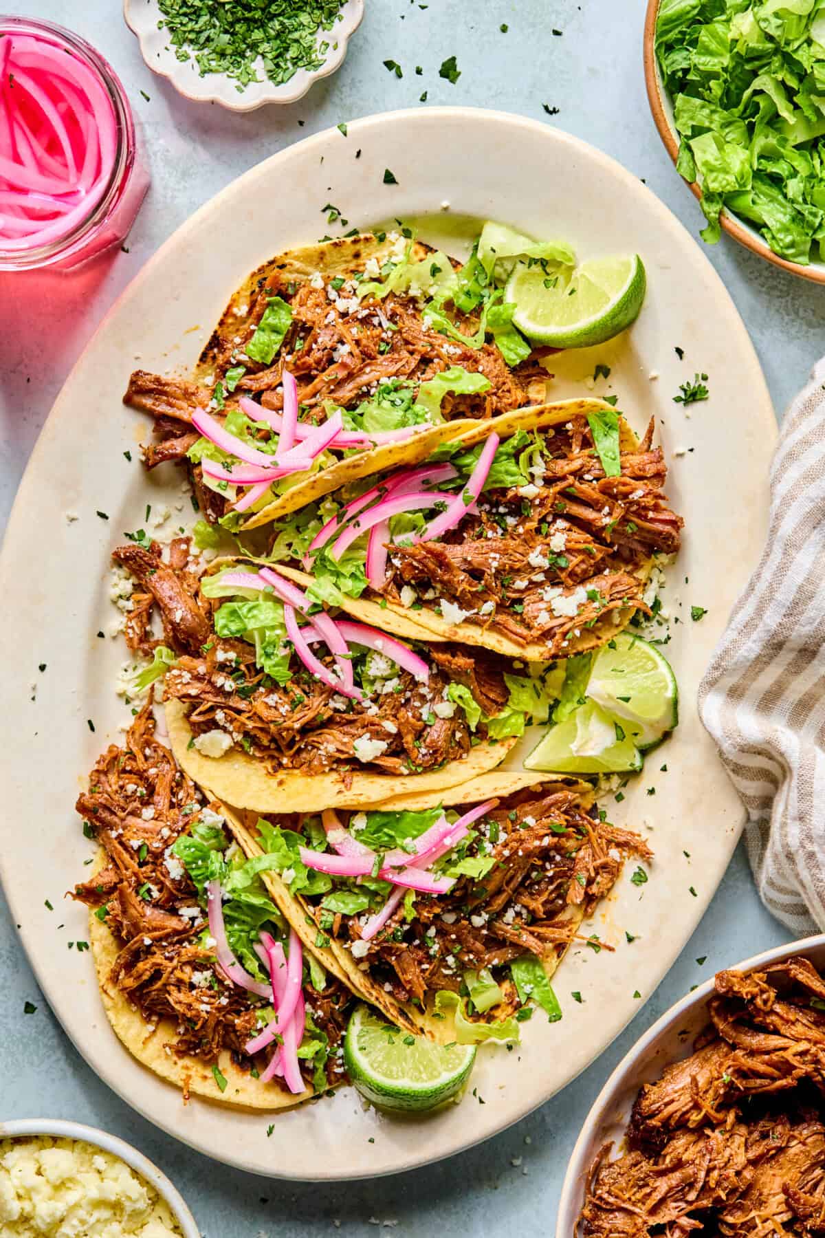 Three pulled beef tacos topped with lettuce, pickled onions, cheese, and chopped herbs are served on a platter with lime wedges. Surrounding the platter are bowls of lettuce, herbs, pickled onions, and savory beef.