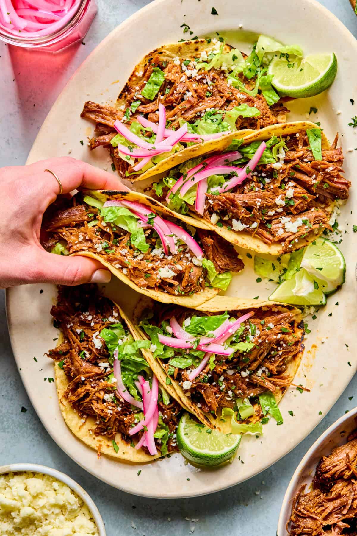 A hand lifts one of four pulled beef tacos on a platter. The tacos are topped with lettuce, pickled red onions, cheese, and served with lime wedges. Other small bowls with food are visible at the edges.