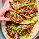 A hand lifts one of four pulled beef tacos on a platter. The tacos are topped with lettuce, pickled red onions, cheese, and served with lime wedges. Other small bowls with food are visible at the edges.