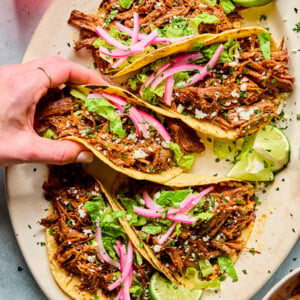 A hand lifts one of four pulled beef tacos on a platter. The tacos are topped with lettuce, pickled red onions, cheese, and served with lime wedges. Other small bowls with food are visible at the edges.