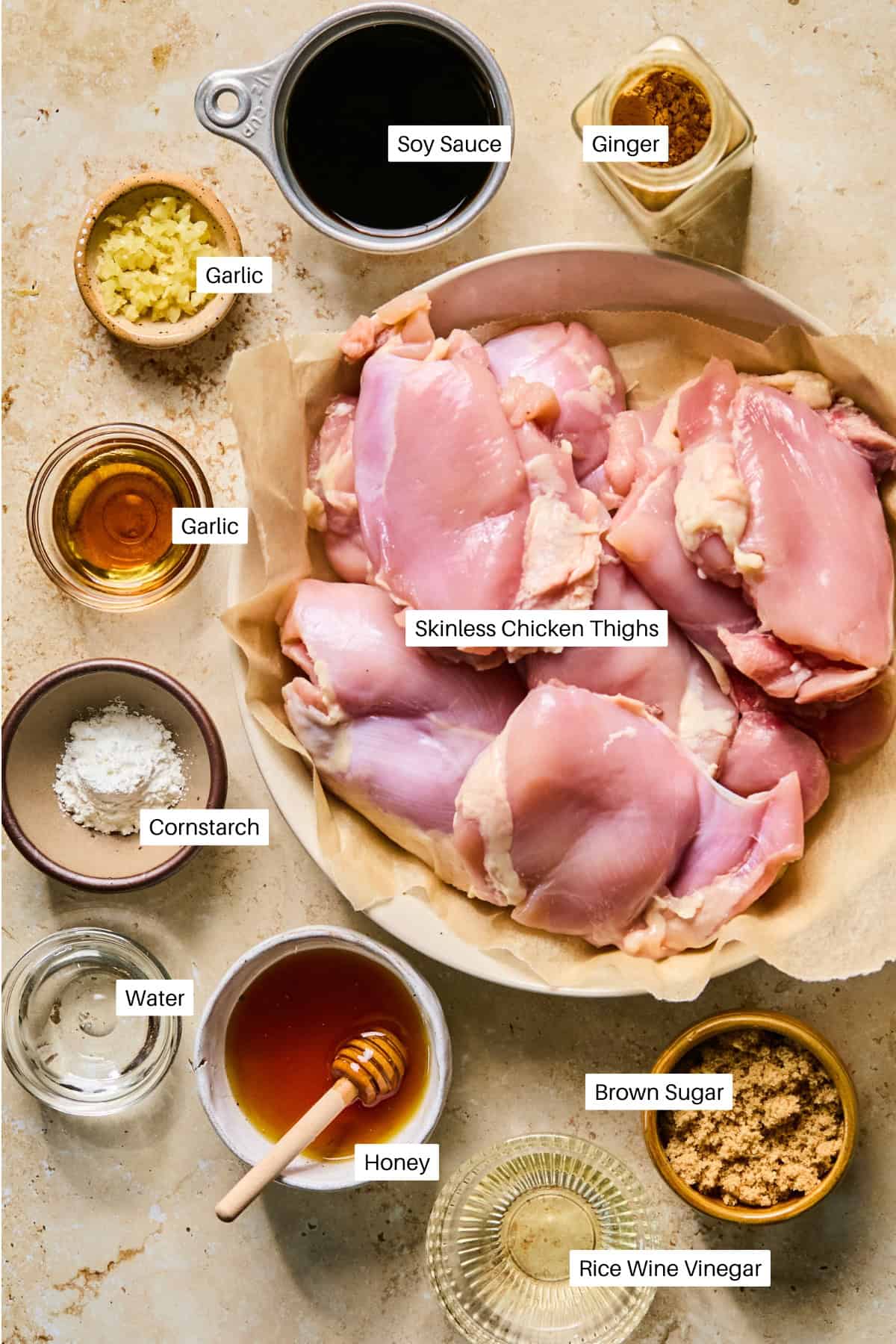 Top-down view of labeled ingredients for air fryer teriyaki chicken thighs: skinless chicken thighs, soy sauce, ginger, garlic, cornstarch, water, honey, brown sugar, and rice wine vinegar, all neatly arranged on a beige countertop.