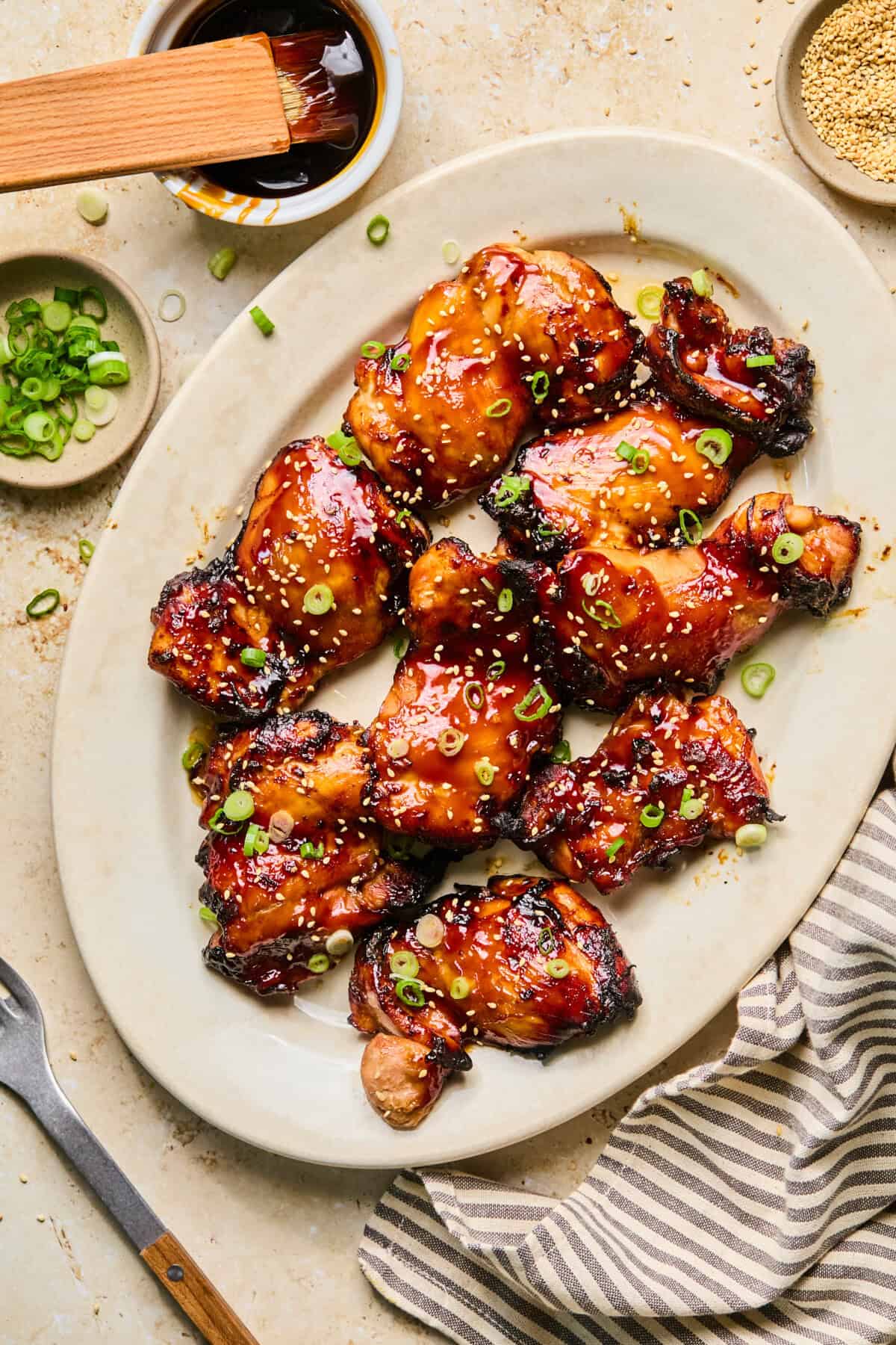 Air fryer teriyaki chicken thighs are grilled and glazed, then topped with chopped green onions on an oval plate. A bowl of sauce, sesame seeds, green onions, and a striped napkin are arranged nearby for added flavor and presentation.