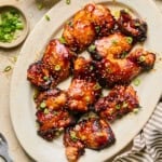 Air fryer teriyaki chicken thighs are grilled and glazed, then topped with chopped green onions on an oval plate. A bowl of sauce, sesame seeds, green onions, and a striped napkin are arranged nearby for added flavor and presentation.