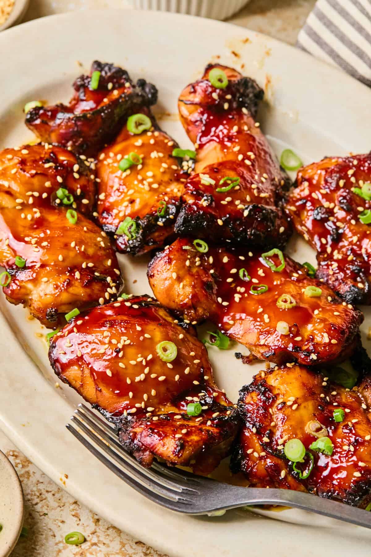 Air fryer teriyaki chicken thighs, glazed to perfection and topped with sesame seeds and chopped green onions, are arranged on a white plate with a fork beside them.