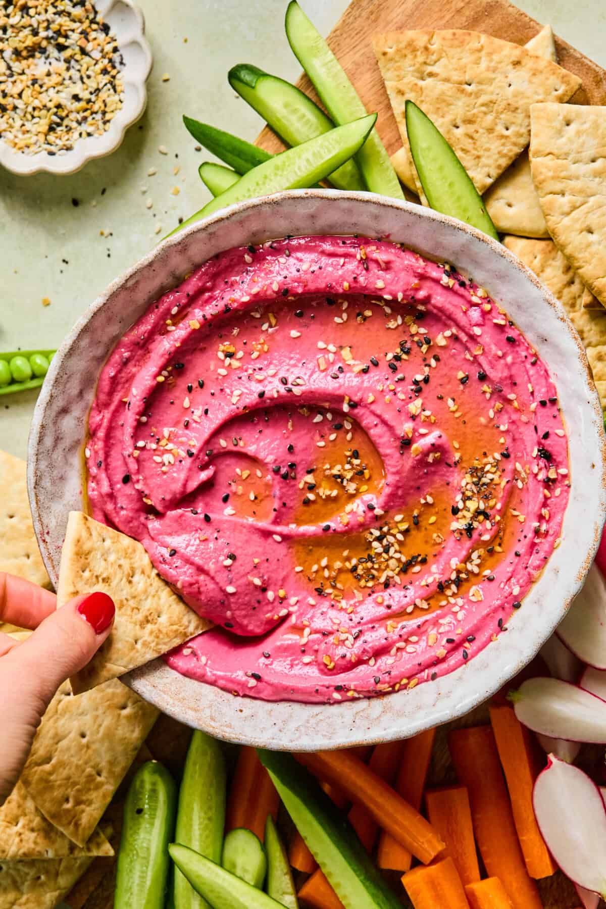 A bowl of bright pink beetroot hummus topped with seeds and oil, surrounded by pita bread, carrot sticks, cucumber slices, and a hand dipping pita into the vibrant hummus.