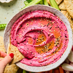 A bowl of bright pink beetroot hummus topped with seeds and oil, surrounded by pita bread, carrot sticks, cucumber slices, and a hand dipping pita into the vibrant hummus.