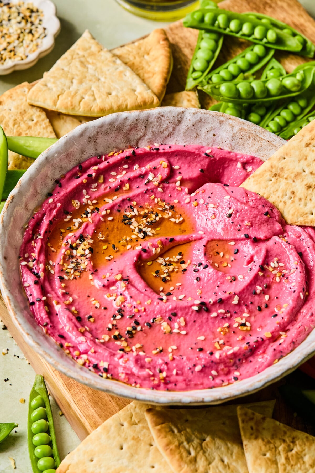 A bowl of vibrant pink beetroot hummus drizzled with oil and sprinkled with seeds, surrounded by pita chips and fresh snap peas on a wooden board.