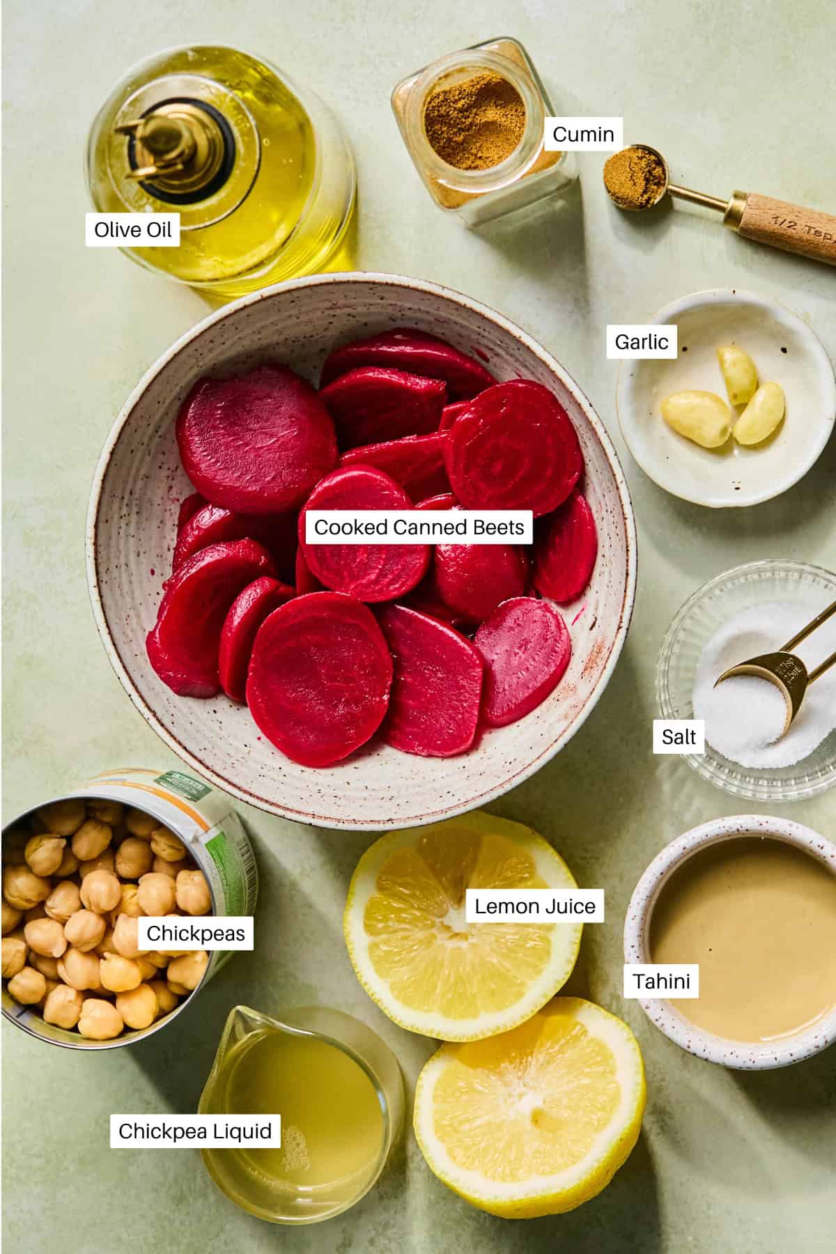Labeled ingredients for beetroot hummus: bowl of cooked canned beets, olive oil, cumin, garlic cloves, salt, chickpeas, lemon juice, chickpea liquid, and tahini arranged on a light green surface.