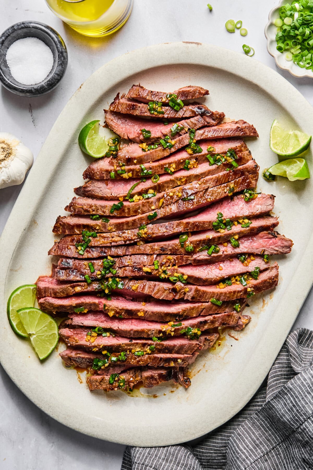 Sliced grilled steak, marinated in a flavorful flank steak marinade, is arranged on a platter, topped with chopped herbs and minced garlic, with lime wedges on the side. A bowl of salt, garlic bulb, and a small dish of green onions are nearby.