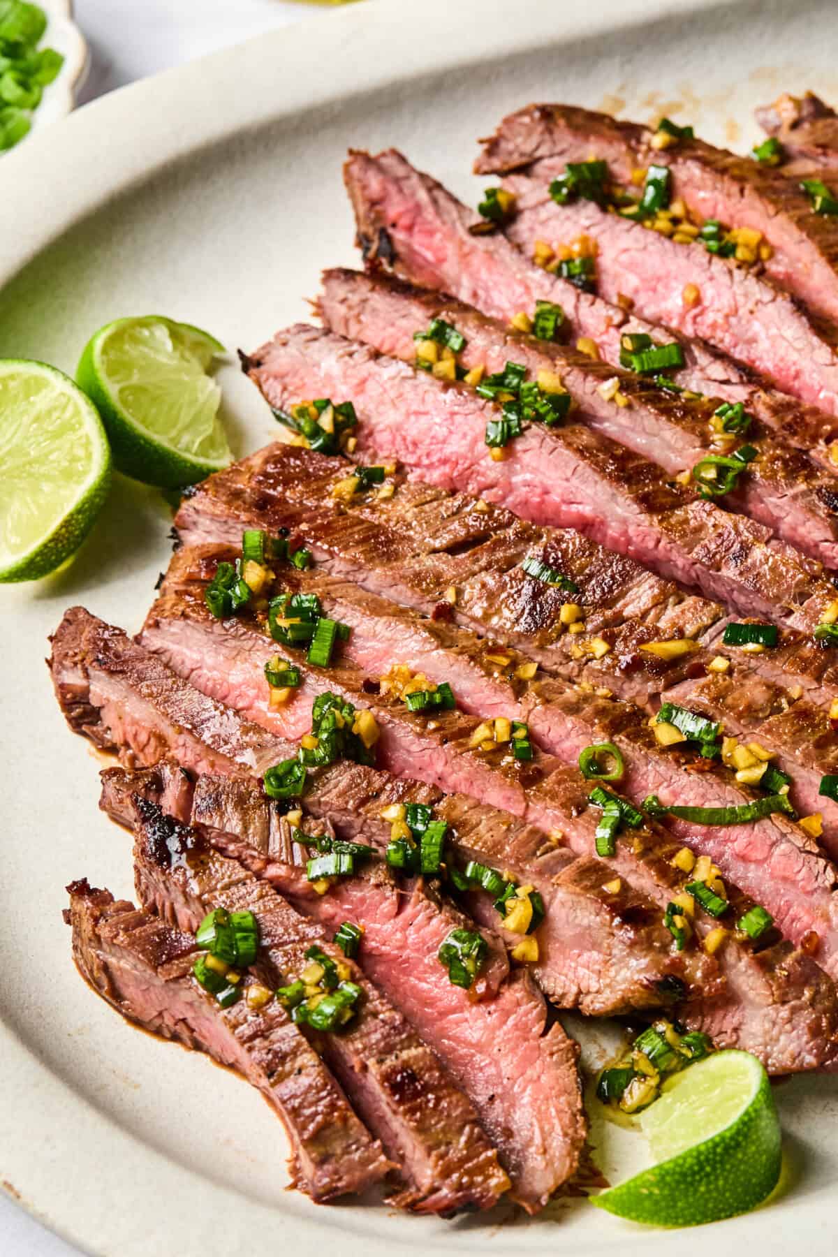 Sliced medium-rare steak, marinated in a flavorful Flank Steak Marinade and topped with chopped herbs and garlic, served on a plate with lime wedges on the side.