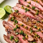 Sliced medium-rare steak, marinated in a flavorful Flank Steak Marinade and topped with chopped herbs and garlic, served on a plate with lime wedges on the side.