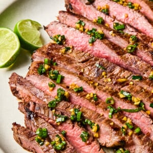Sliced medium-rare steak, marinated in a flavorful Flank Steak Marinade and topped with chopped herbs and garlic, served on a plate with lime wedges on the side.