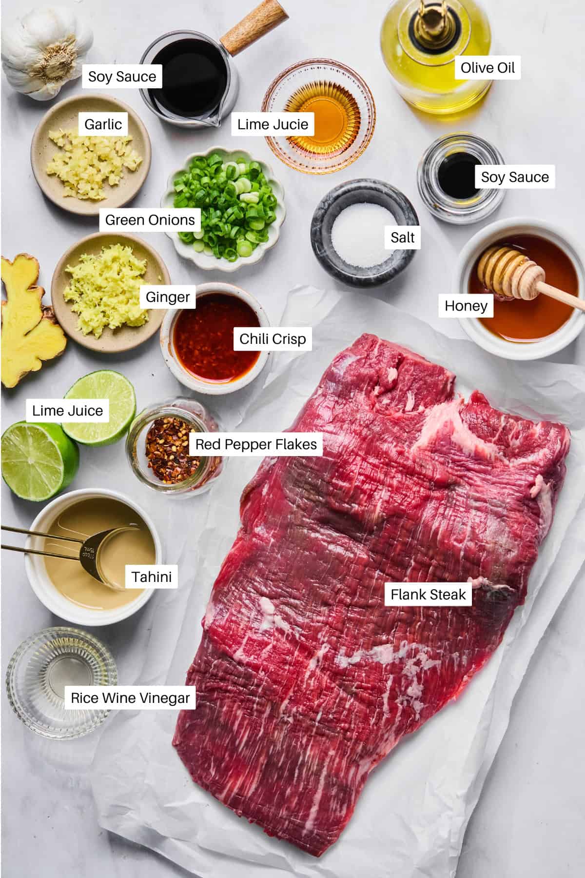 Raw flank steak on parchment surrounded by labeled bowls of soy sauce, olive oil, garlic, lime juice, green onions, salt, honey, ginger, chili crisp, red pepper flakes, tahini, and rice wine vinegar—everything you need for a flavorful Flank Steak Marinade.