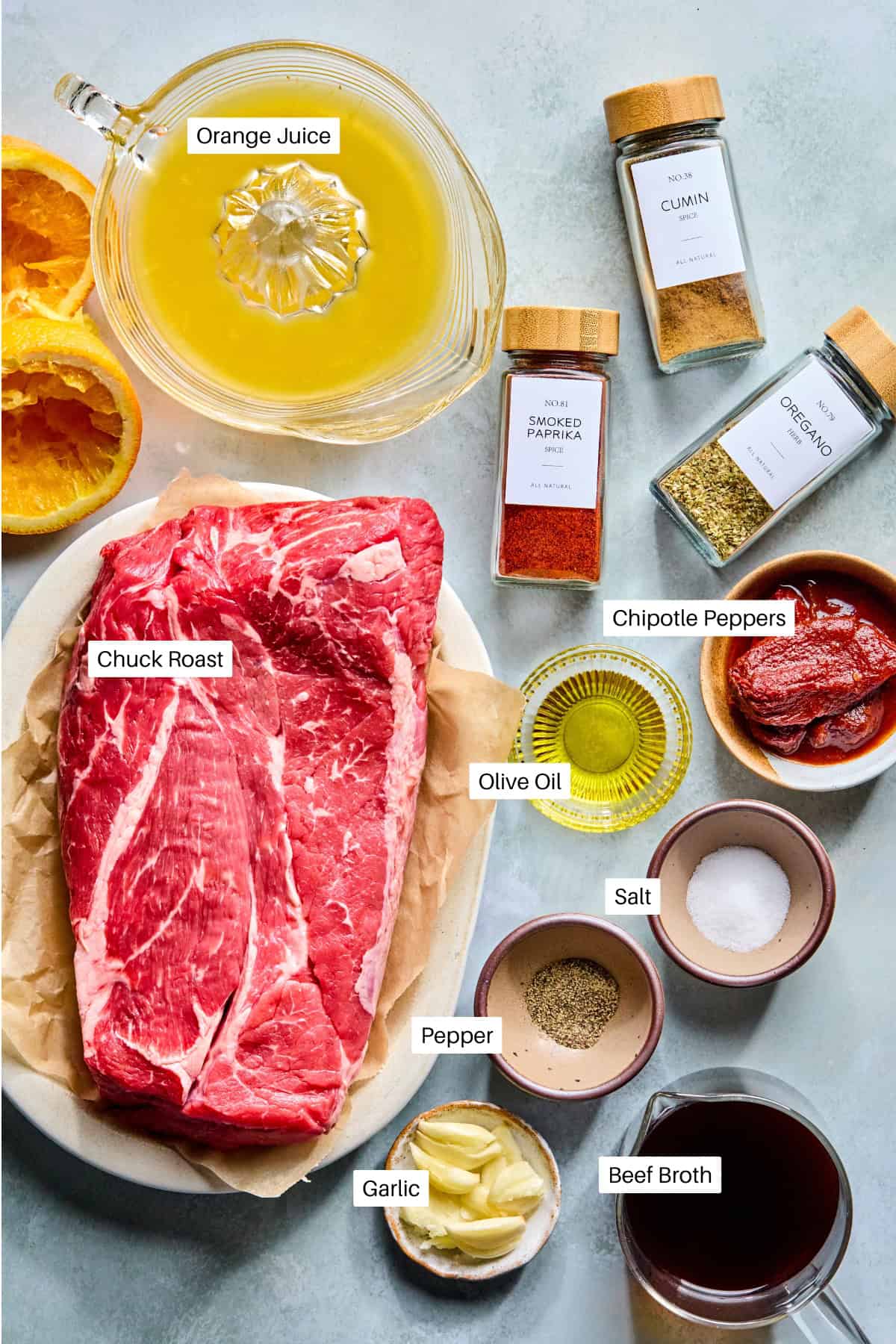 A flat lay of ingredients for Pulled Beef, featuring chuck roast, sliced garlic, chipotle peppers, orange juice, olive oil, beef broth, salt, pepper, cumin, smoked paprika, and oregano—all arranged on a light surface.