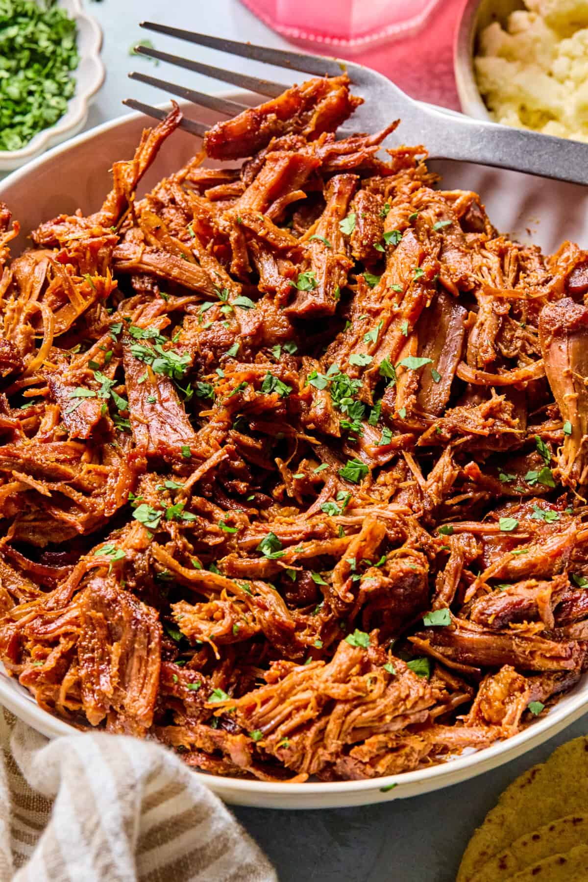 A bowl filled with shredded, seasoned pulled beef garnished with chopped fresh herbs, with a fork resting on the edge. Sides of mashed potatoes and chopped green herbs are partially visible nearby.