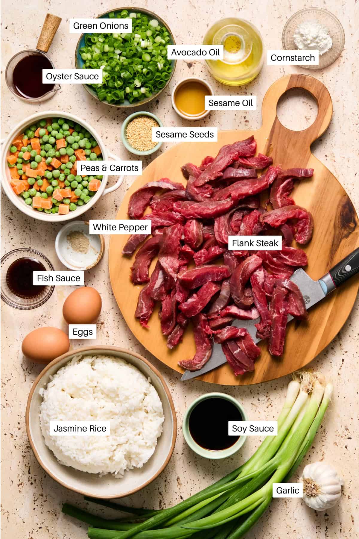Sliced flank steak on a wooden board with knife, surrounded by bowls of rice, eggs, seasonings, sauces, chopped green onions, peas and carrots, oil, garlic, and sesame seeds—each ingredient labeled for the ultimate steal fried rice experience.