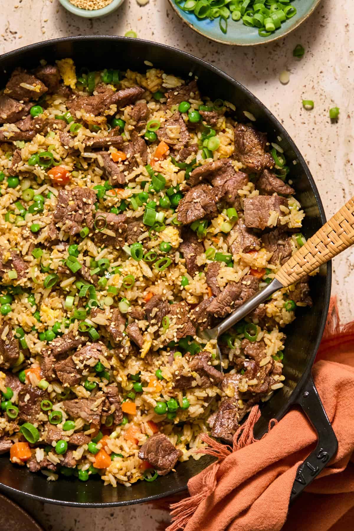 A skillet filled with beef steal fried rice, mixed with peas, carrots, and scrambled eggs, garnished with green onions. A spoon rests in the skillet with chopped green onions and a napkin nearby.