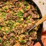 A skillet filled with beef steal fried rice, mixed with peas, carrots, and scrambled eggs, garnished with green onions. A spoon rests in the skillet with chopped green onions and a napkin nearby.
