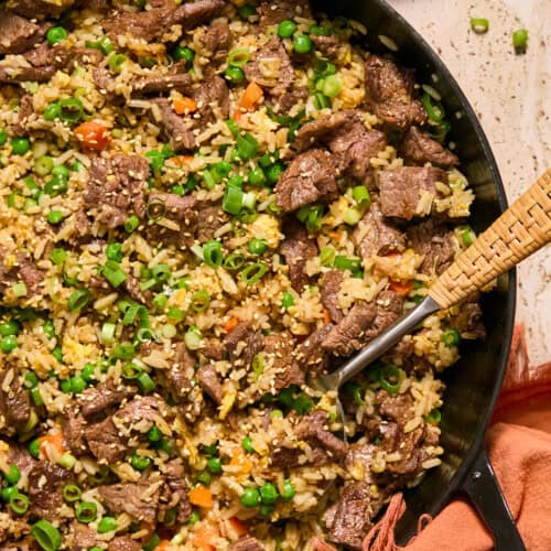 A skillet filled with beef steal fried rice, mixed with peas, carrots, and scrambled eggs, garnished with green onions. A spoon rests in the skillet with chopped green onions and a napkin nearby.