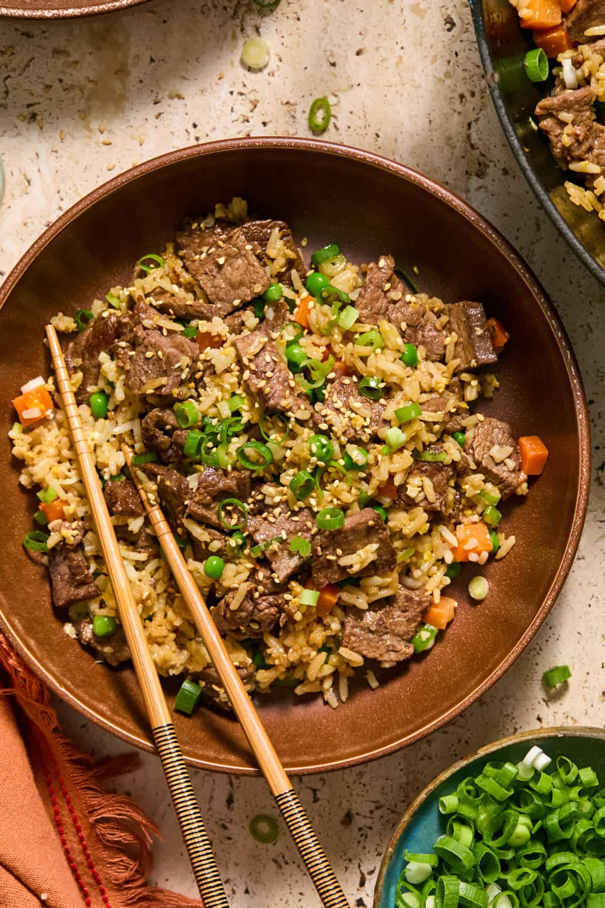 A bowl of beef steal fried rice with chopped green onions, carrots, and sesame seeds, served with a pair of chopsticks resting on the bowl.