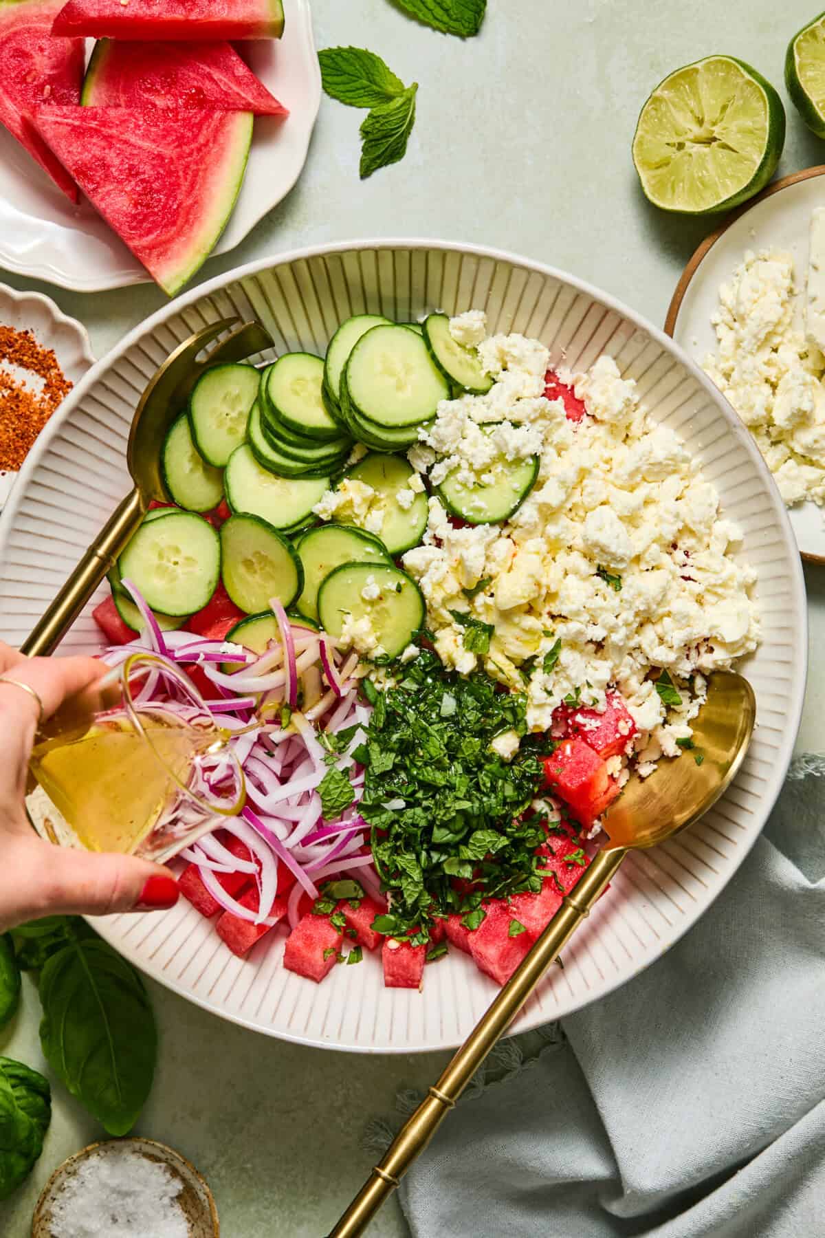 A hand pours dressing onto a vibrant watermelon feta salad with sliced cucumber, red onion, chopped herbs, and crumbled cheese in a white bowl. Slices of lime and fresh watermelon are nearby.