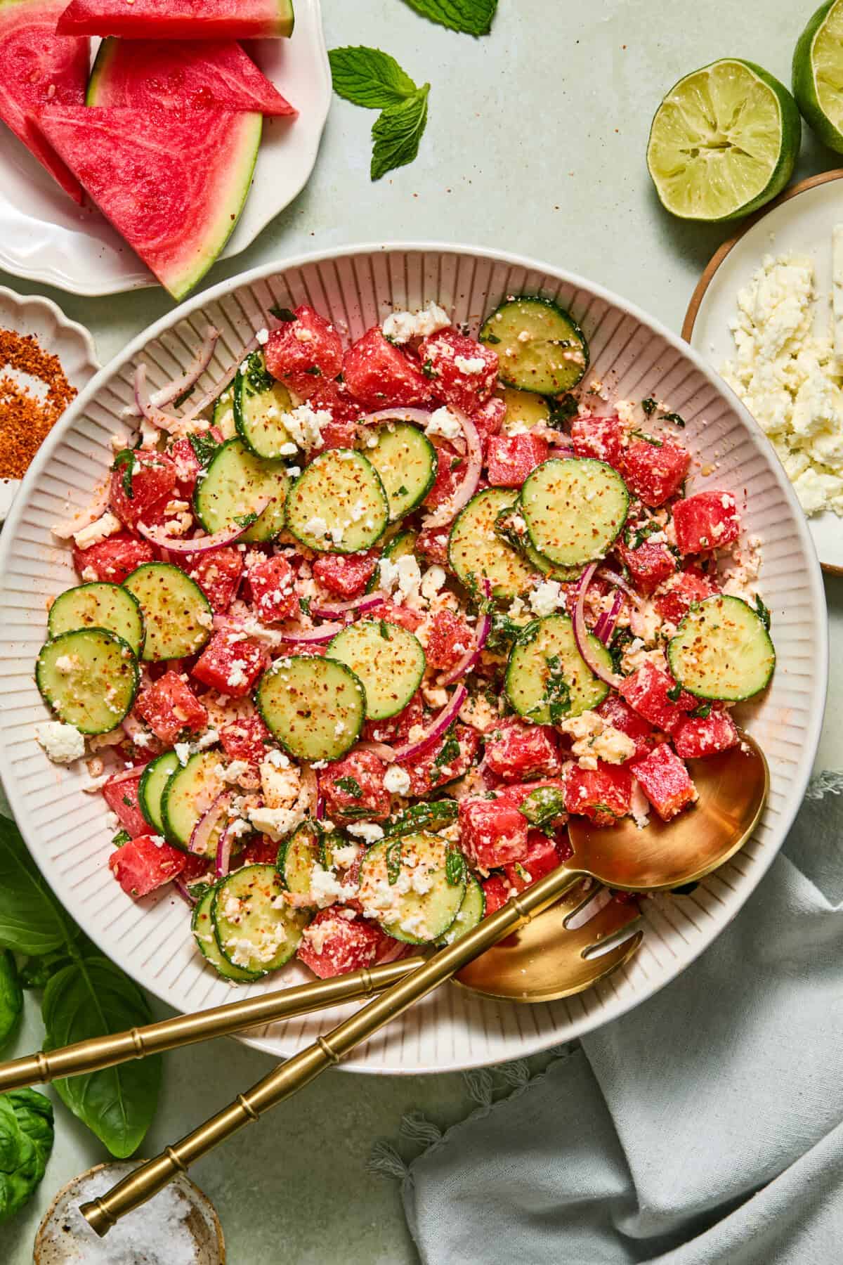 A vibrant watermelon feta salad sits in a white bowl with gold utensils, featuring refreshing watermelon, crisp cucumber, red onion, and creamy feta cheese. Nearby on a light table are lime halves, extra watermelon slices, and chunks of feta for garnishing.