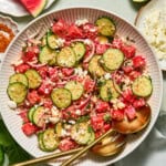 A vibrant watermelon feta salad sits in a white bowl with gold utensils, featuring refreshing watermelon, crisp cucumber, red onion, and creamy feta cheese. Nearby on a light table are lime halves, extra watermelon slices, and chunks of feta for garnishing.