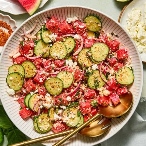 A vibrant watermelon feta salad sits in a white bowl with gold utensils, featuring refreshing watermelon, crisp cucumber, red onion, and creamy feta cheese. Nearby on a light table are lime halves, extra watermelon slices, and chunks of feta for garnishing.