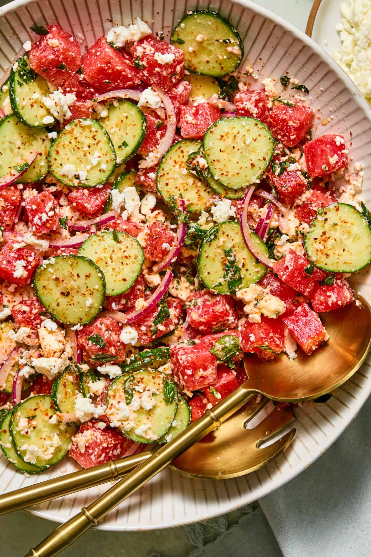 A close-up of a refreshing watermelon feta salad with sliced cucumbers, watermelon cubes, red onion, feta cheese, herbs, and spices, served in a white bowl with gold serving utensils.