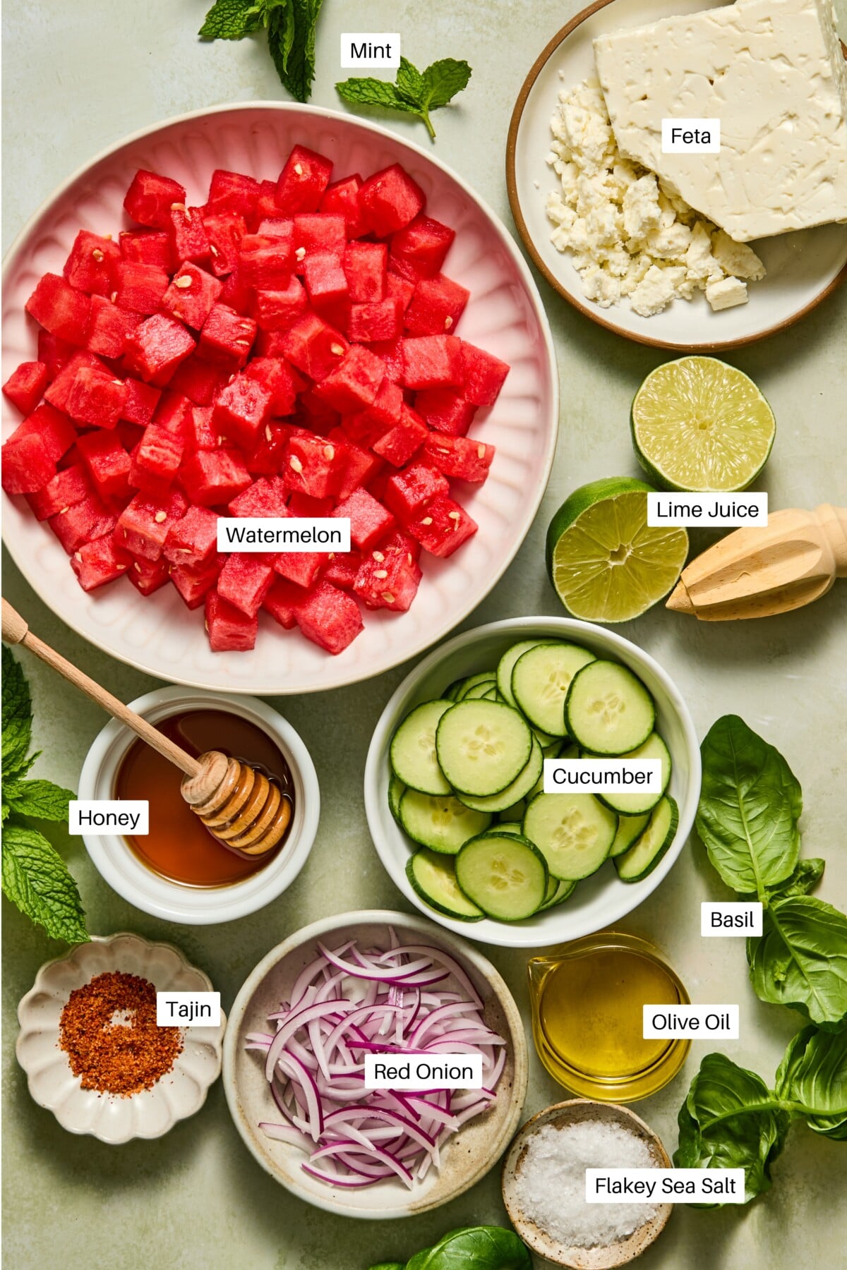 A flat lay of ingredients for a refreshing watermelon feta salad, featuring cubed watermelon, creamy feta, lime juice, cucumber slices, red onion, honey, olive oil, Tajin seasoning, basil, mint, and flakey sea salt.