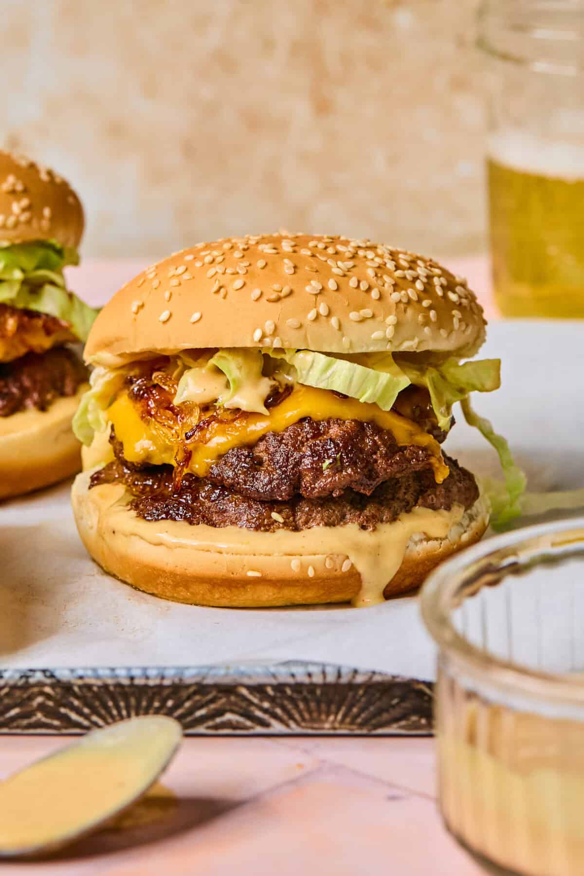 A cheeseburger with lettuce, beetroot hummus, and sauce on a sesame bun sits on parchment paper, with another burger, a dish of sauce, and a glass of beer in the background.