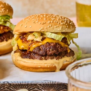 A cheeseburger with lettuce, beetroot hummus, and sauce on a sesame bun sits on parchment paper, with another burger, a dish of sauce, and a glass of beer in the background.