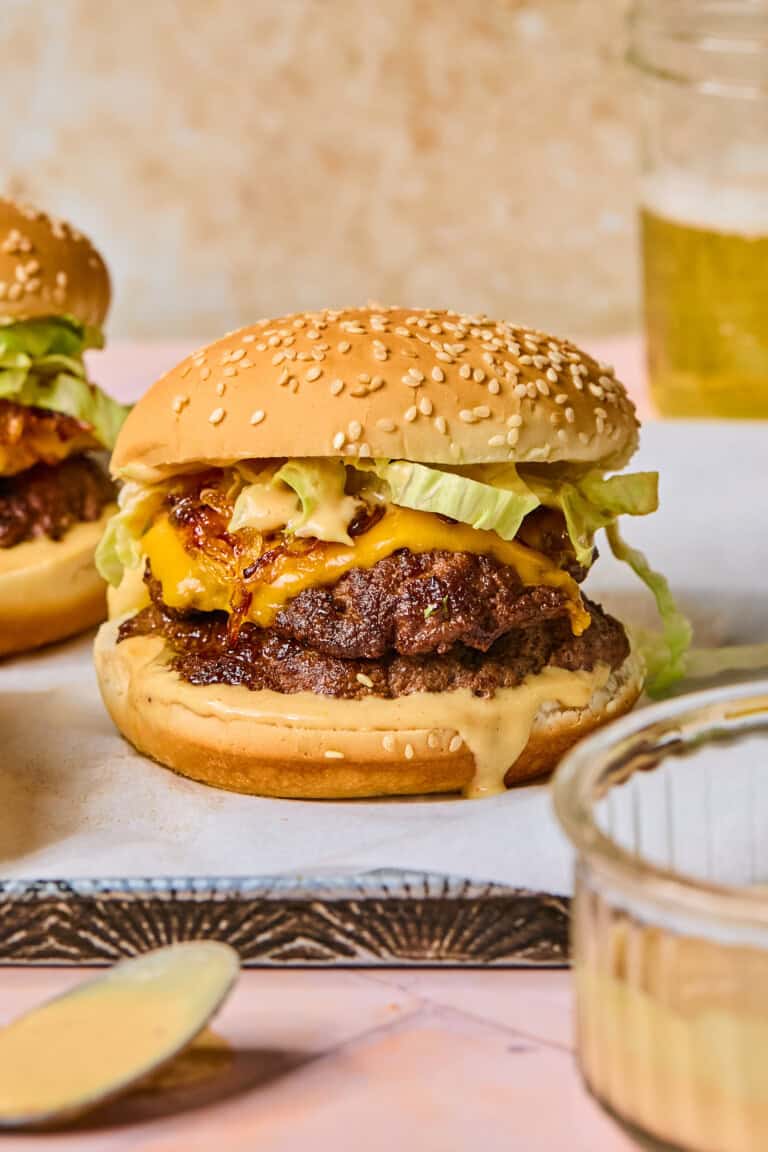 A cheeseburger with lettuce, beetroot hummus, and sauce on a sesame bun sits on parchment paper, with another burger, a dish of sauce, and a glass of beer in the background.