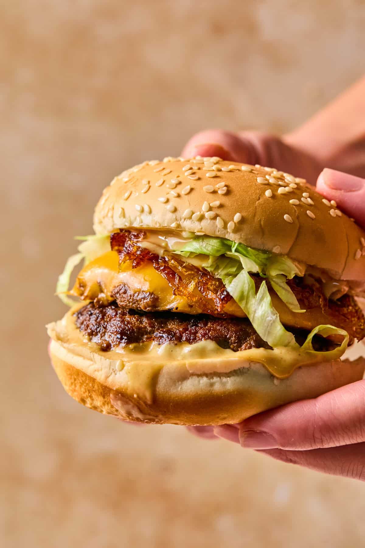 A hand holds a sesame seed bun burger with a beef patty, melted cheese, crispy bacon, shredded lettuce, and a dollop of beetroot hummus sauce, against a blurred brown background.