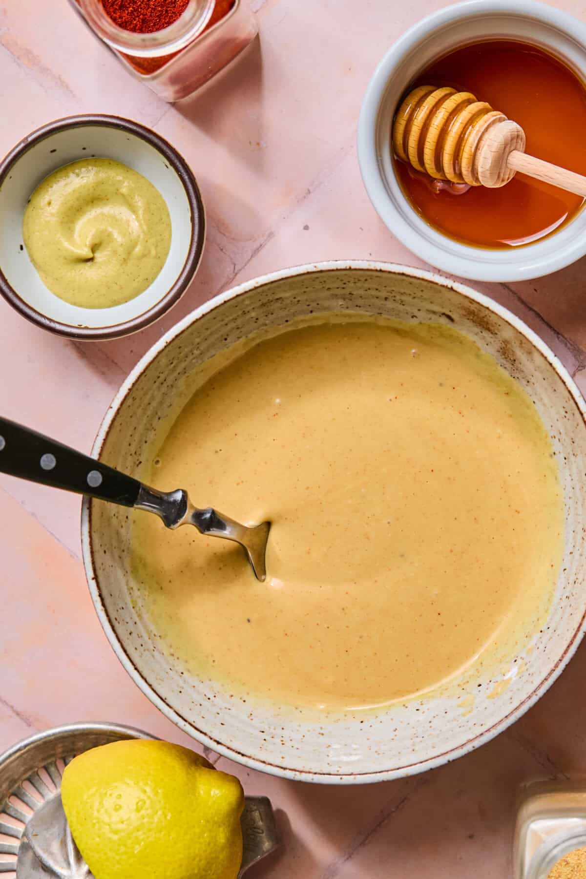A bowl of honey mustard sauce with a spoon, surrounded by a lemon, honey jar, mustard, and a small dish of paprika on a pink surface—perfect for those craving a tasty copy-cat Chick-fil-A sauce experience at home.