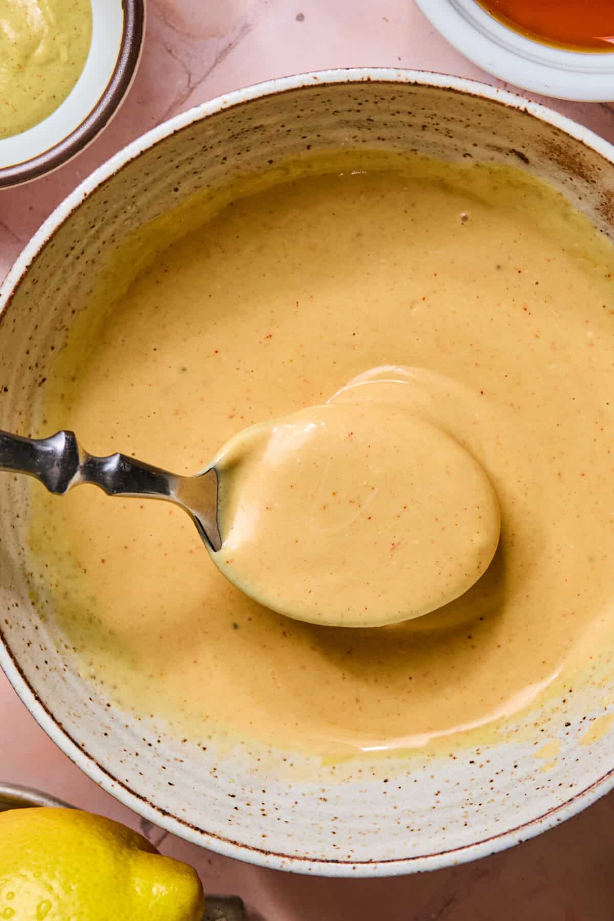 A spoon holding creamy, light brown copy-cat Chick-fil-A sauce above a speckled ceramic bowl, with a lemon and small dishes partially visible nearby on a pink surface.