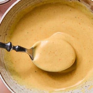 A spoon holding creamy, light brown copy-cat Chick-fil-A sauce above a speckled ceramic bowl, with a lemon and small dishes partially visible nearby on a pink surface.