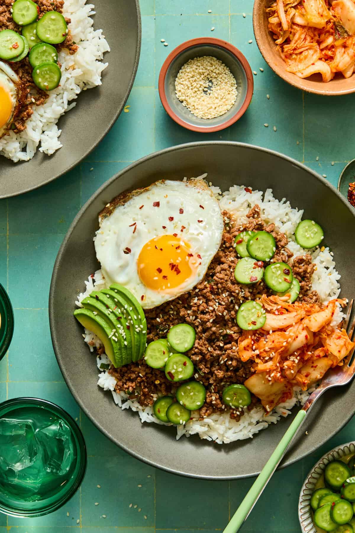 A bowl of rice topped with ground meat, a sunny-side-up egg, sliced cucumbers, avocado, kimchi, and sprinkled with sesame seeds sits on a green table next to water glasses, more bowls, and a refreshing watermelon feta salad.