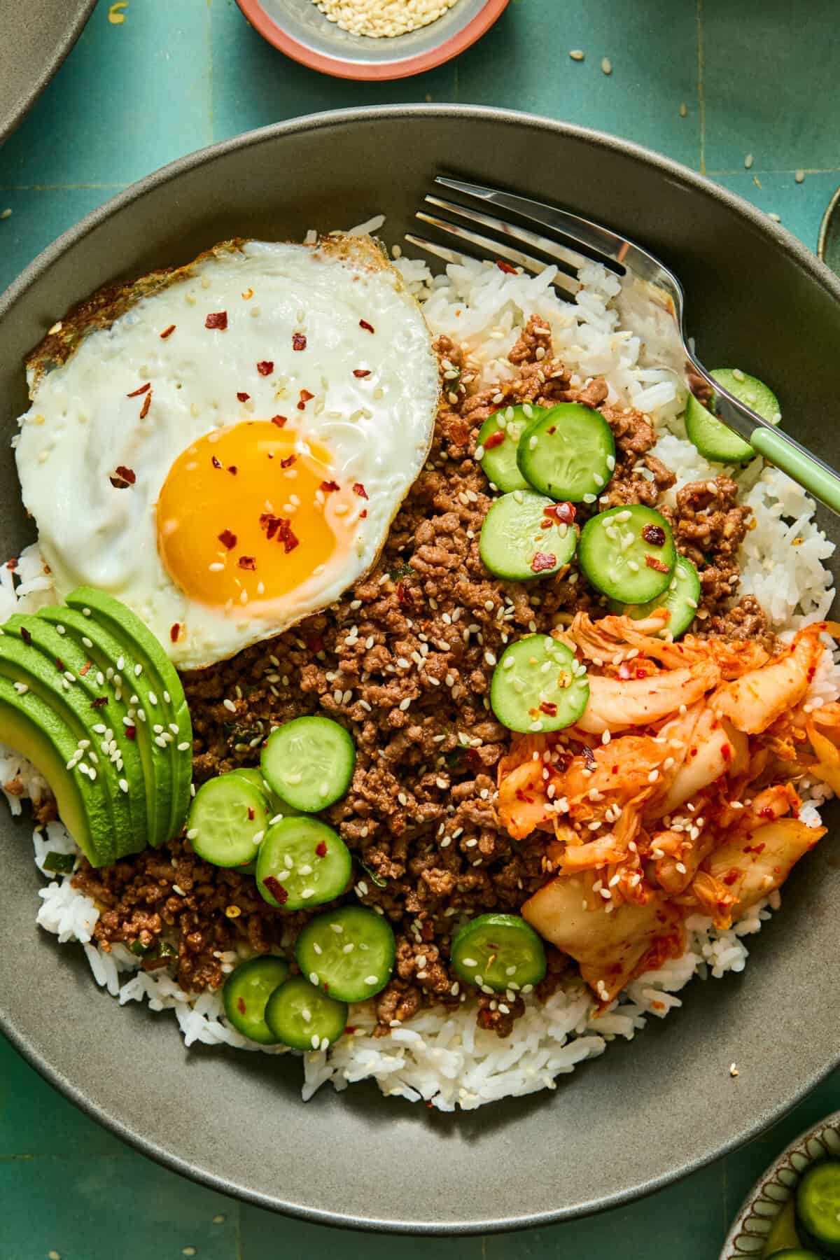 A bowl of white rice topped with ground beef, sliced cucumber, avocado, kimchi, and a sunny-side-up egg, garnished with sesame seeds and red pepper flakes—perfectly paired with a refreshing watermelon feta salad for a vibrant and satisfying meal.