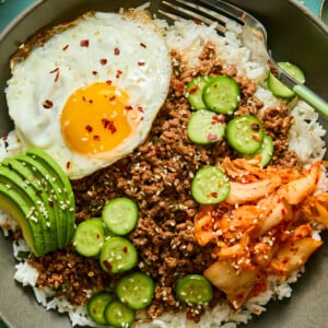 A bowl of white rice topped with ground beef, sliced cucumber, avocado, kimchi, and a sunny-side-up egg, garnished with sesame seeds and red pepper flakes—perfectly paired with a refreshing watermelon feta salad for a vibrant and satisfying meal.