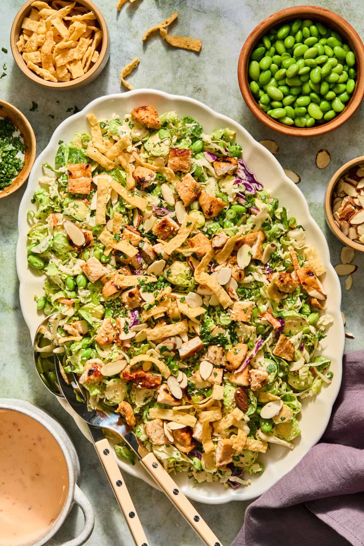 Large oval platter of chopped salad with grilled chicken, sliced almonds, crispy wonton strips, and green edamame. Surrounding bowls hold extra toppings, including a vibrant watermelon feta salad. A cup of pink dressing and utensils are beside the platter.