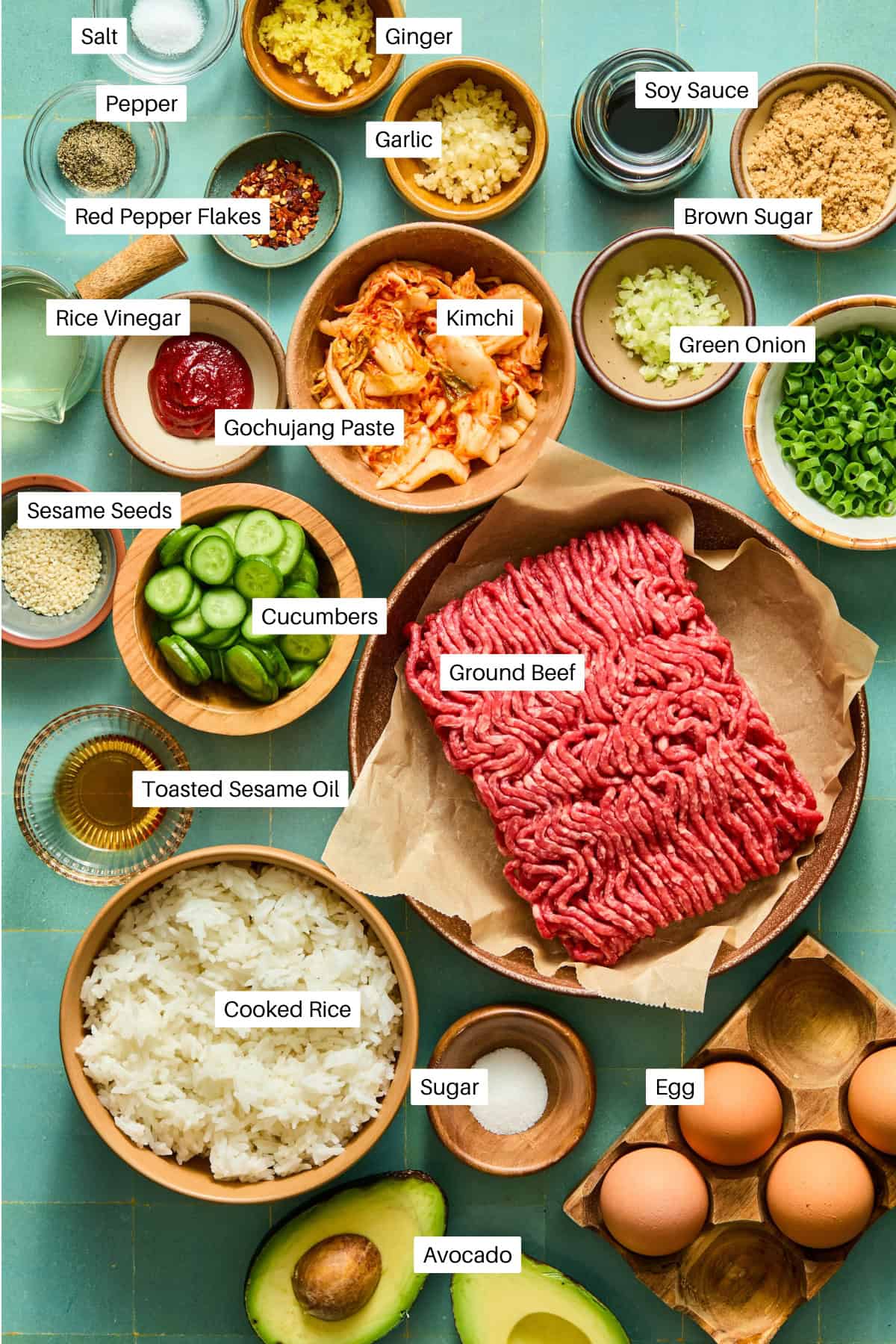 A top-down view of neatly arranged ingredients for a Korean-inspired rice bowl, including ground beef, kimchi, cooked rice, avocado, eggs, seasonings, saucy condiments, vegetables, spices, and sesame seeds on a blue surface—perfectly complemented by the vibrant freshness of a watermelon feta salad nearby.